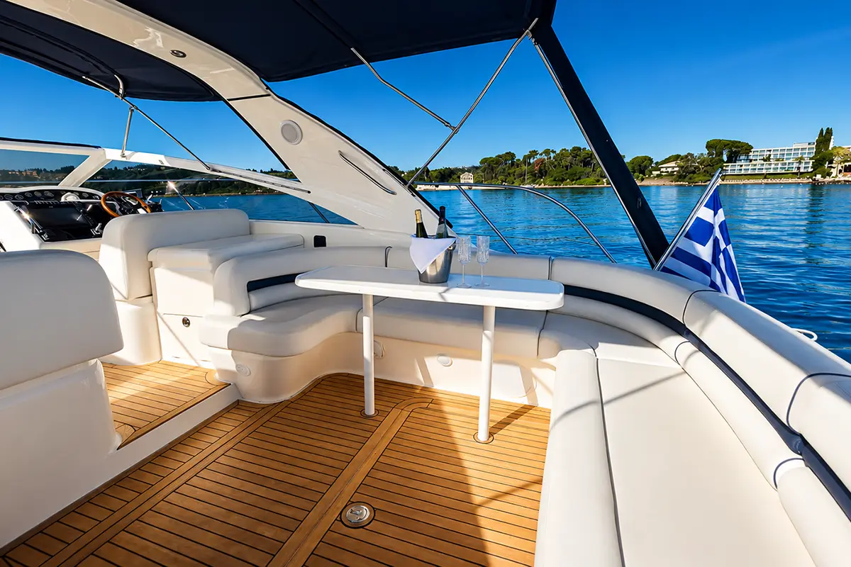Luxury yacht cockpit with white curved seating, a small table set with champagne and flutes, on a teak deck, calm blue water and Greek flag in view.