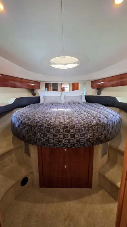 Round bed with a blue patterned duvet in a yacht cabin, surrounded by built-in seating and wood cabinetry.