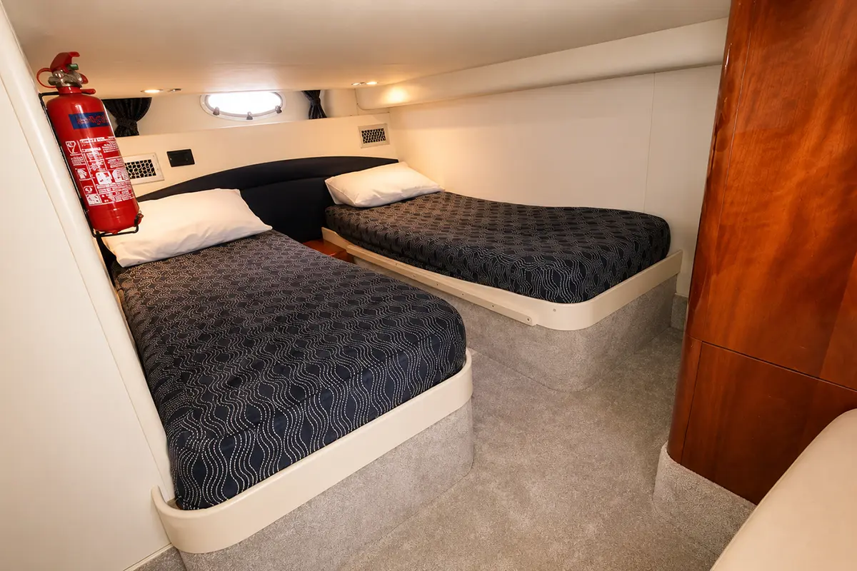 Twin-bed cabin with navy patterned bedspreads, porthole window, and a red fire extinguisher mounted on the wall nearby.